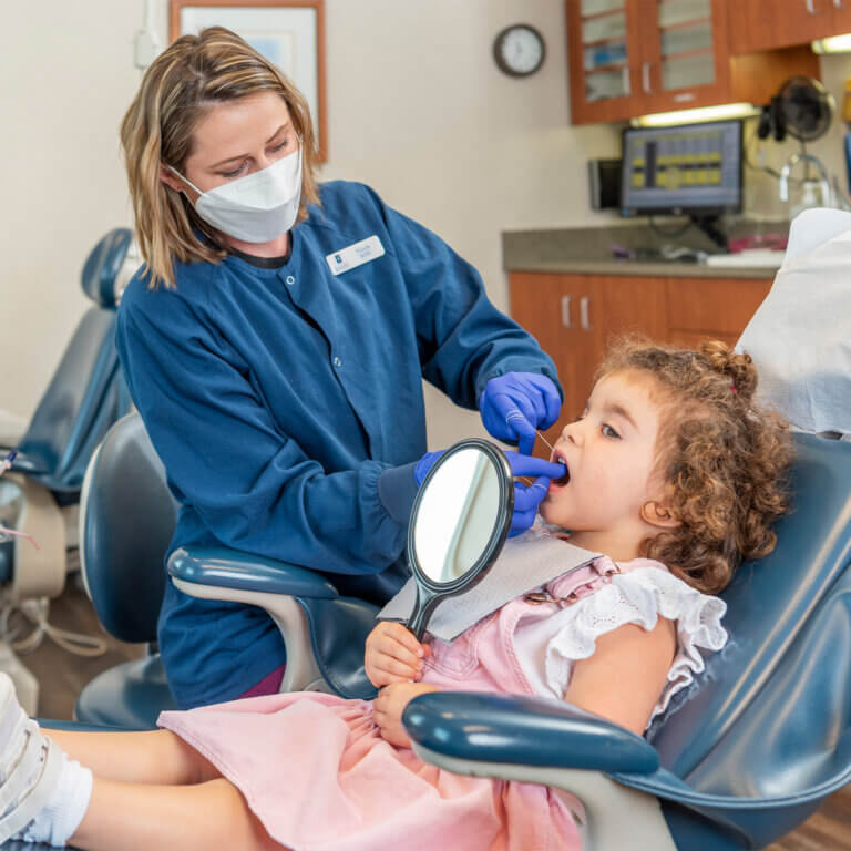 Masked dental assistant flossing a toddler's teeth